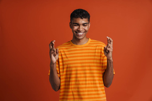 Young black man with piercing holding fingers crossed for good luck isolated over red background - Valokuva, kuva