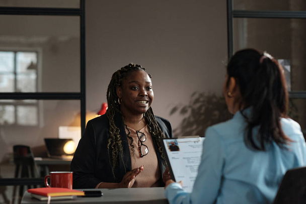 Afrikaanse jonge vrouw praten over haar werkervaring naar interviewer terwijl ze zitten aan tafel tijdens sollicitatiegesprek - Foto, afbeelding