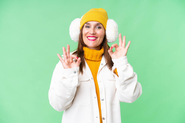 Mujer de mediana edad con manguitos de invierno sobre fondo de croma clave aislado que muestra un signo de ok con los dedos - Foto, Imagen