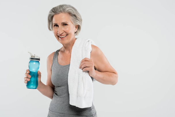 ευχαριστημένη ηλικιωμένη γυναίκα με λευκή πετσέτα στον ώμο κρατώντας αθλητικό μπουκάλι με νερό που απομονώνεται σε γκρι  - Φωτογραφία, εικόνα