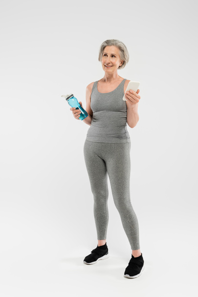 full length of happy ηλικιωμένη γυναίκα στα αθλητικά κρατώντας αθλητικό μπουκάλι με νερό και smartphone σε γκρι  - Φωτογραφία, εικόνα
