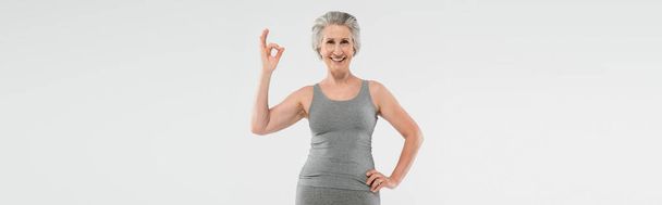 mujer alegre y jubilada en ropa deportiva posando con la mano en la cadera y mostrando bien aislado en gris, bandera  - Foto, Imagen