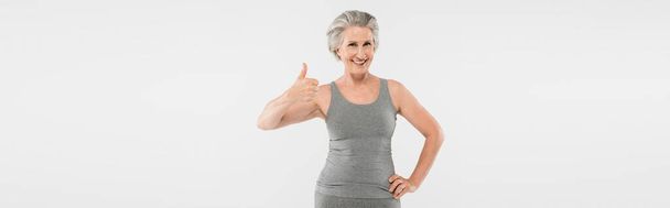 cheerful and retired woman in sportswear posing with hand on hip and showing like isolated on grey, banner  - Фото, изображение