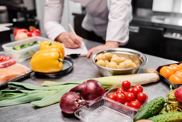 Close-up of fresh vegetables on table in commercial kitchen with chef making menu in background - Photo, Image