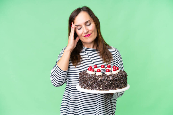 Orta yaşlı, beyaz bir kadın, doğum günü pastasını izole edilmiş krom anahtar zemininde tutuyor ve baş ağrısı var. - Fotoğraf, Görsel