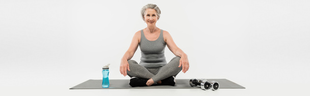 longitud completa de la mujer mayor feliz sentada en postura del yoga en la estera cerca de las mancuernas y de la botella del deporte en gris, bandera  - Foto, imagen