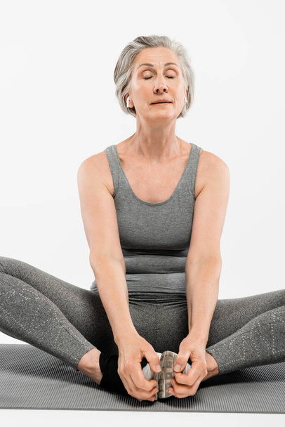 Seniorin mit drahtlosen Kopfhörern sitzt in Yoga-Pose auf Matte isoliert auf grau  - Foto, Bild