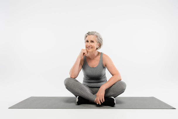 piena lunghezza della donna anziana soddisfatto in auricolari wireless ascoltare musica e seduto in posa yoga sul grigio  - Foto, immagini