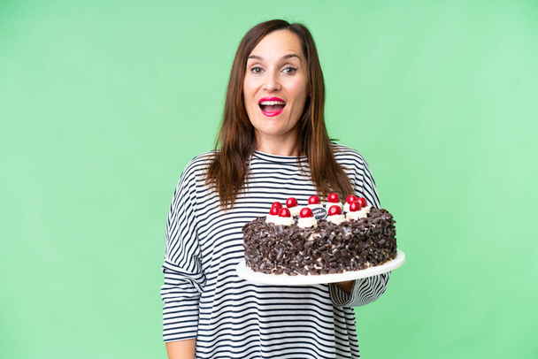 Середньовічна біла жінка тримає торт на день народження над ізольованим ключовим фоном хрому з несподіваним виразом обличчя
 - Фото, зображення