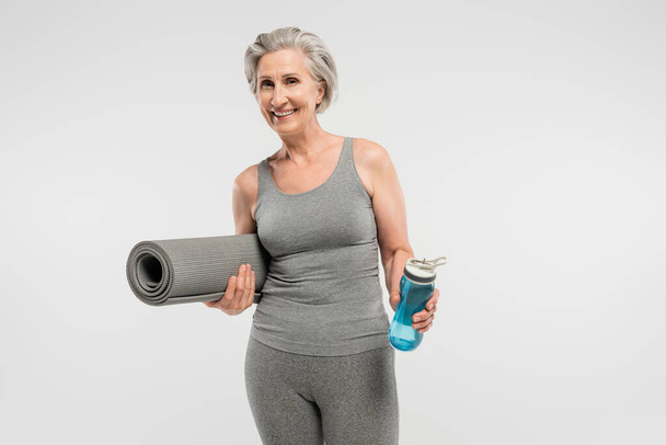 χαρούμενη ηλικιωμένη γυναίκα κρατώντας στρώμα γυμναστικής και αθλητικό μπουκάλι με νερό που απομονώνεται σε γκρι  - Φωτογραφία, εικόνα