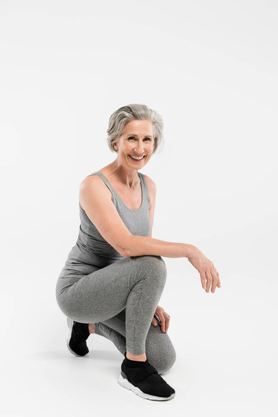 volle Länge der fröhlichen Seniorin in Sportbekleidung lächelnd, während sie auf grau sitzt  - Foto, Bild