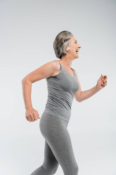side view of cheerful senior woman with grey hair running isolated on grey  - Fotografie, Obrázek