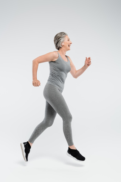 full length of happy senior woman with grey hair running on grey background  - Photo, Image