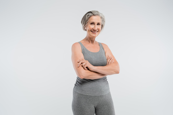 cheerful senior woman in sportswear standing with folded arms isolated on grey  - Foto, Imagen