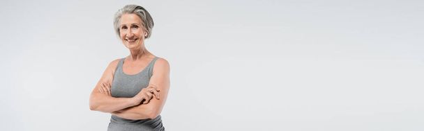 carefree senior woman in sportswear standing with folded arms isolated on grey, banner  - Foto, afbeelding