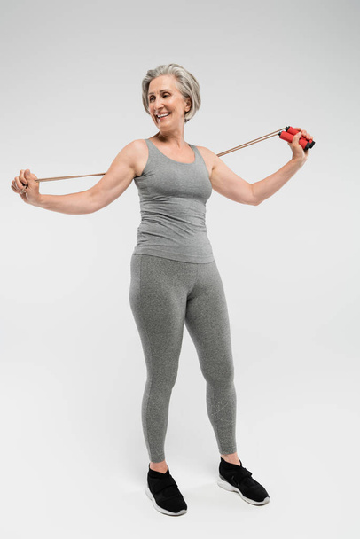 full length of cheerful senior woman holding skipping rope on grey  - 写真・画像