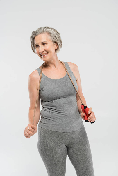 pleased senior woman with grey hair holding skipping rope isolated on grey  - Фото, зображення