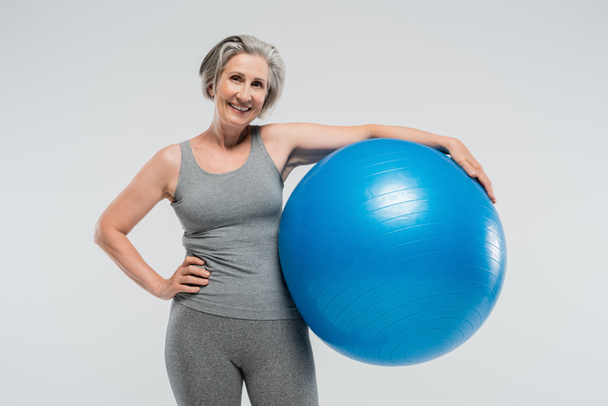 fröhliche Seniorin in Sportkleidung hält blauen Fitnessball isoliert auf grau  - Foto, Bild