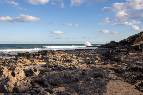 Połączenie piasku i skalistego wybrzeża na plaży Oceanu Atlantyckiego. Niebieskie niebo z białymi chmurami. Parque Natural Dunas de Corralejo, Fuerteventura, Hiszpania. - Zdjęcie, obraz