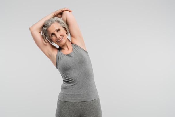 Spor kıyafetleri içinde neşeli bir büyükanne, gri renkte izole edilmiş bir şekilde egzersiz yapıyor ve gülümsüyor.  - Fotoğraf, Görsel