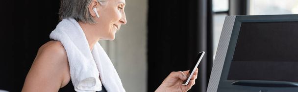 szczęśliwy starszy kobieta w bezprzewodowy słuchawki gospodarstwa smartfon stojąc w pobliżu bieżni w siłowni, baner  - Zdjęcie, obraz