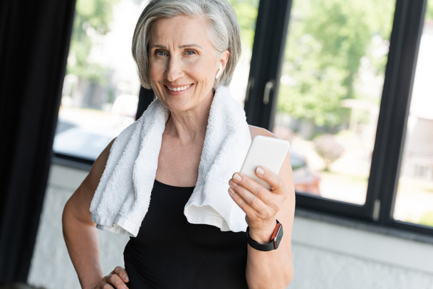 veselá starší žena s ručníkem na ramenou pomocí smartphonu při poslechu hudby v bezdrátovém sluchátku  - Fotografie, Obrázek