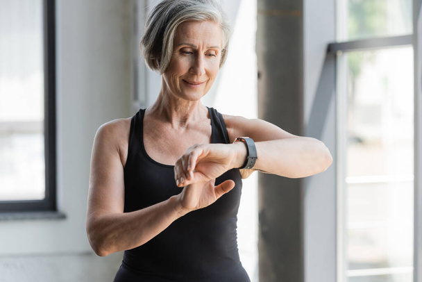 happy senior woman in black tank top checking activity on fitness tracker in gym  - 写真・画像
