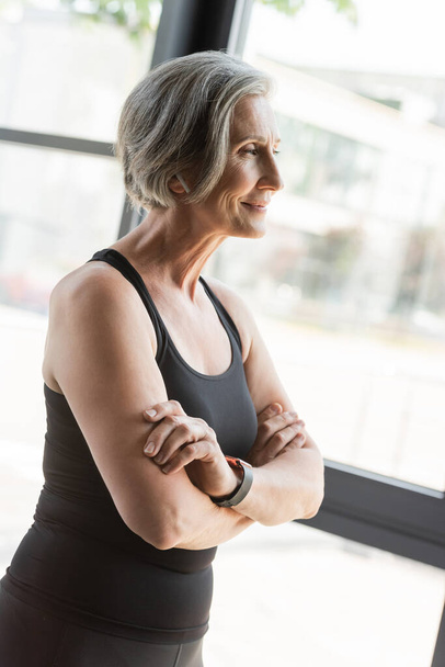 cheerful senior sportswoman in tank top smiling while standing with crossed arms in gym  - Photo, Image