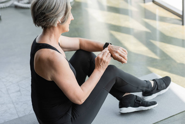 fit starší žena s šedými vlasy sedí a pomocí fitness tracker po cvičení v tělocvičně  - Fotografie, Obrázek