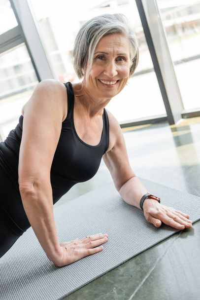 senior sportswoman smiling while exercising on fitness mat in gym  - 写真・画像