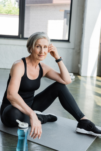 senior woman in sportswear listening music and sitting on fitness mat in gym  - Photo, Image