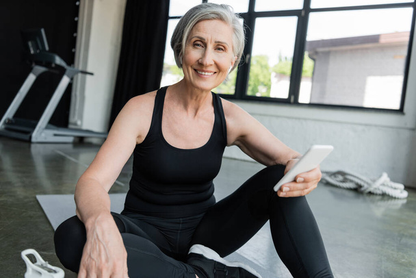 cheerful sportswoman using mobile phone and sitting on fitness mat in gym  - Photo, Image