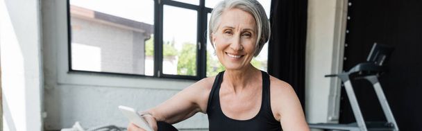 cheerful sportswoman with grey hair using mobile phone after working out in gym, banner - Foto, Imagen