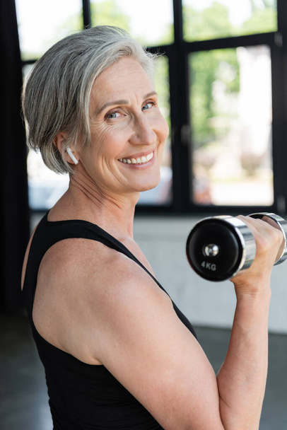 overjoyed senior woman in wireless earphone working out with dumbbell in gym  - Foto, Imagen
