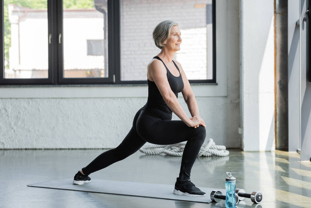 full length of cheerful senior woman in sportswear doing lunges on fitness mat  - Foto, Imagen