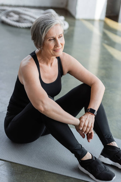 full length of happy senior woman in black leggings and tank top sitting on fitness mat  - Foto, Imagem