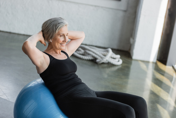 cheerful senior woman in black leggings and tank top exercising on fitness ball  - Zdjęcie, obraz