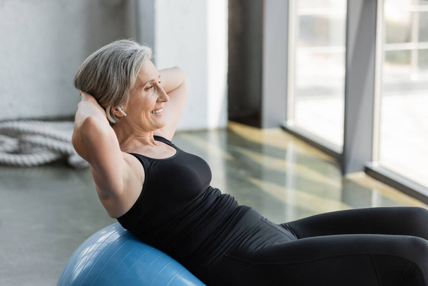 positive senior woman in black leggings and tank top exercising on blue fitness ball  - Fotó, kép