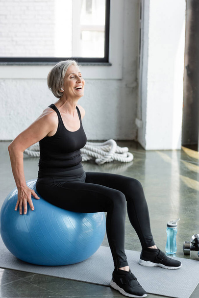 full length of excited senior woman in black leggings and tank top exercising on blue fitness ball  - Photo, Image