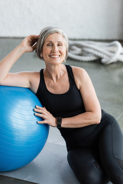 happy senior woman in black leggings and tank top sitting near blue fitness ball in gym  - Foto, afbeelding