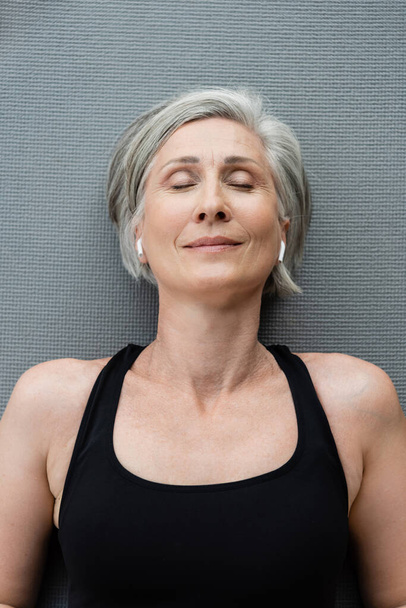 top view of pleased senior woman in wireless earphones lying on fitness mat  - Foto, Bild
