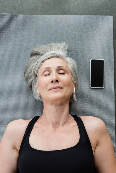 Draufsicht auf glückliche Seniorin in drahtlosen Kopfhörern, die neben Smartphone mit leerem Bildschirm auf Fitnessmatte liegen  - Foto, Bild