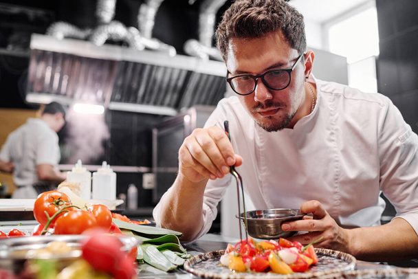 Joven chef en anteojos poniendo ensalada de verduras en plato mientras trabaja en cocina de restaurante - Foto, imagen
