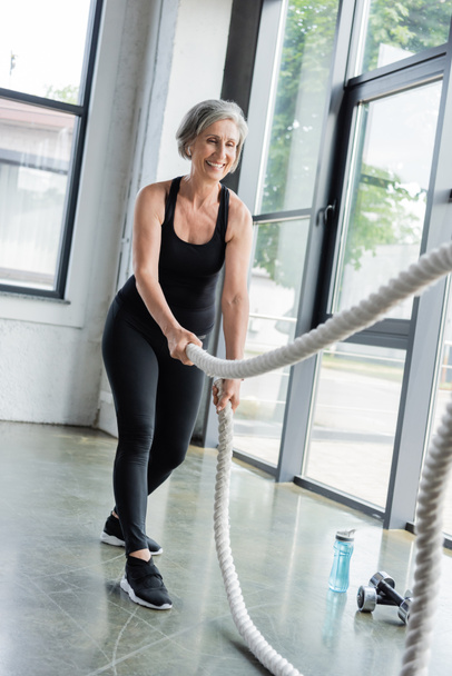 full length of cheerful senior woman working out with battle ropes in gym  - Foto, Imagem