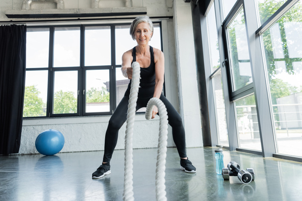 full length of cheerful senior woman training with battle ropes near dumbbells in gym  - Fotó, kép