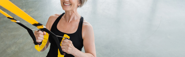 vista cortada de mulher idosa exultante em sportswear preto trabalhando com tiras de suspensão no ginásio, banner  - Foto, Imagem