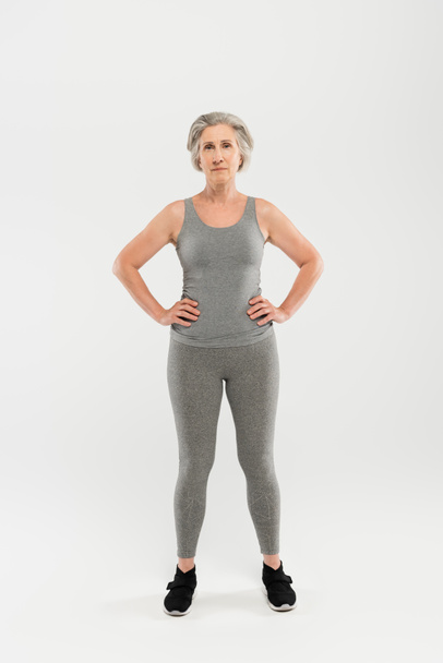 full length of happy and retired woman in sportswear standing with hands on hips on grey - Valokuva, kuva