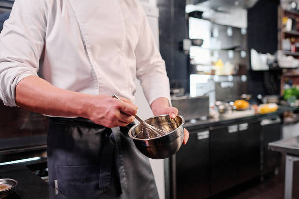 Крупный план шеф-повара в фартуке, взбивающего яйца в миску венчиком во время приготовления пищи на кухне - Фото, изображение