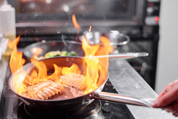 Detailní záběr kuchaře drží pánev nad sporákem a smažení ryb na otevřeném ohni v kuchyni - Fotografie, Obrázek