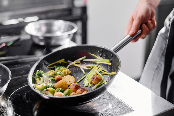 Крупный план профессионального повара, держащего сковороду над плитой и жарящего картофель с овощами - Фото, изображение